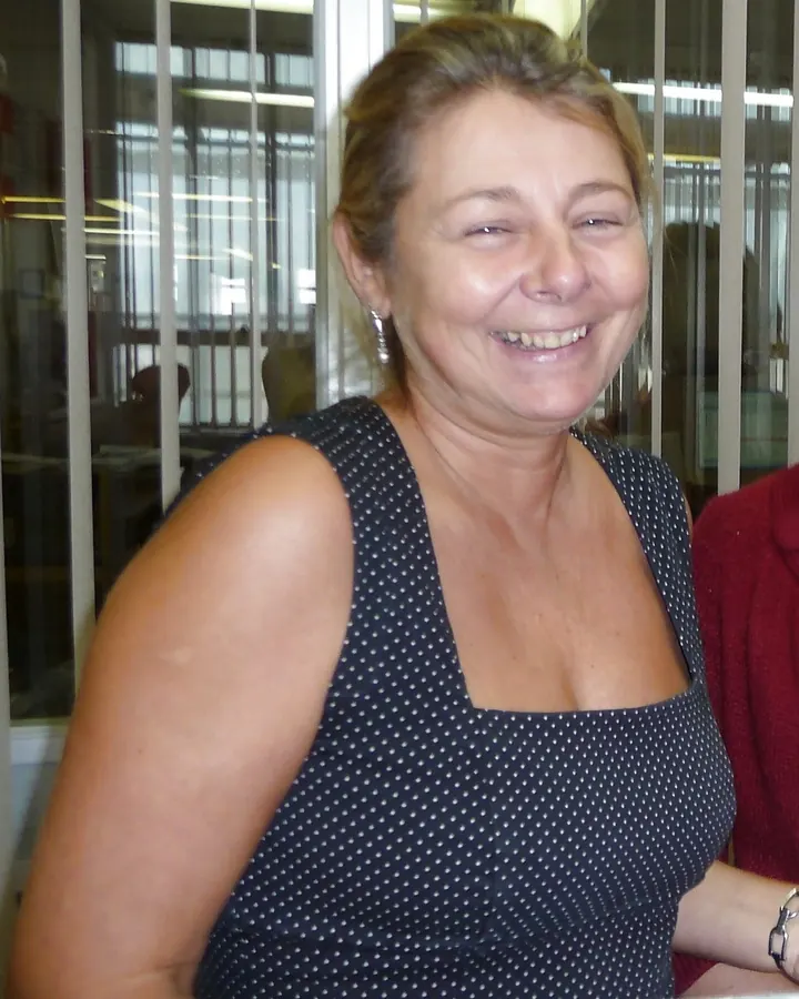 Femme souriante en haut noir à pois blancs, vue de face, devant une structure métallique verticale.