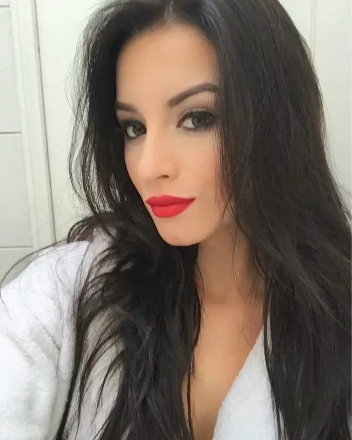 Femme aux cheveux noirs longs et maquillage prononcé, en robe blanche, regardant directement l'appareil photo.