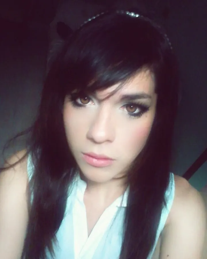 Jeune femme aux cheveux bruns, portant un haut blanc et un foulard noir, posant dans une chambre sombre avec un regard intense.