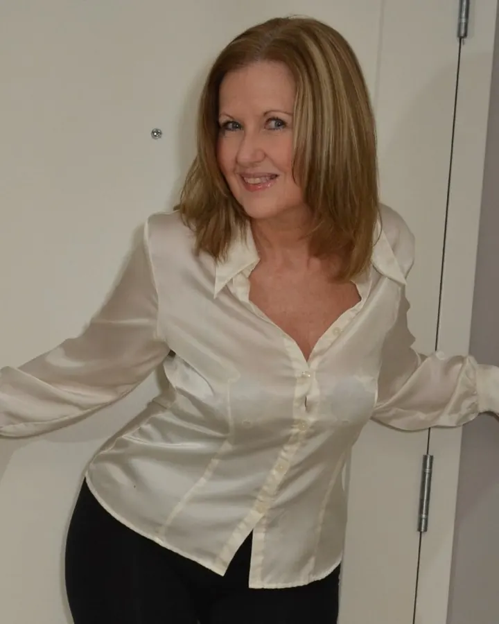 Femme aux cheveux blonds, portant une chemise blanche et une jupe noire, posant près d'une porte blanche avec un sourire.