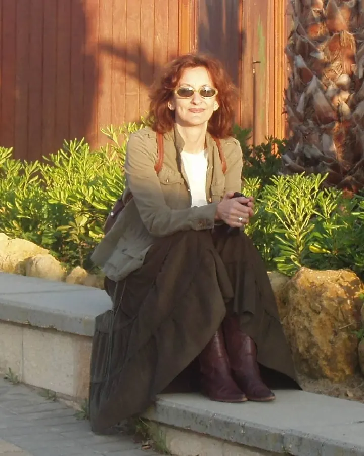 Femme aux cheveux châtains portant des lunettes de soleil, assise sur un muret en extérieur devant une palme et des buissons verts.
